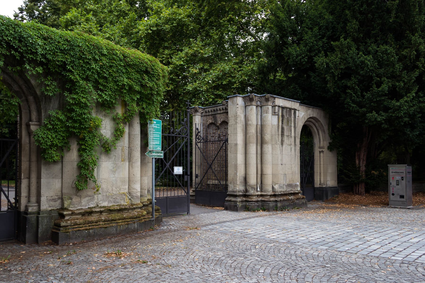 Schrägansicht der steinernen Toranlage an der Kölnstraße mit schmiedeeisernen Gittern, efeubewachsener Bogenpforte und Grabkerzenautomat auf Nordfriedhof Bonn