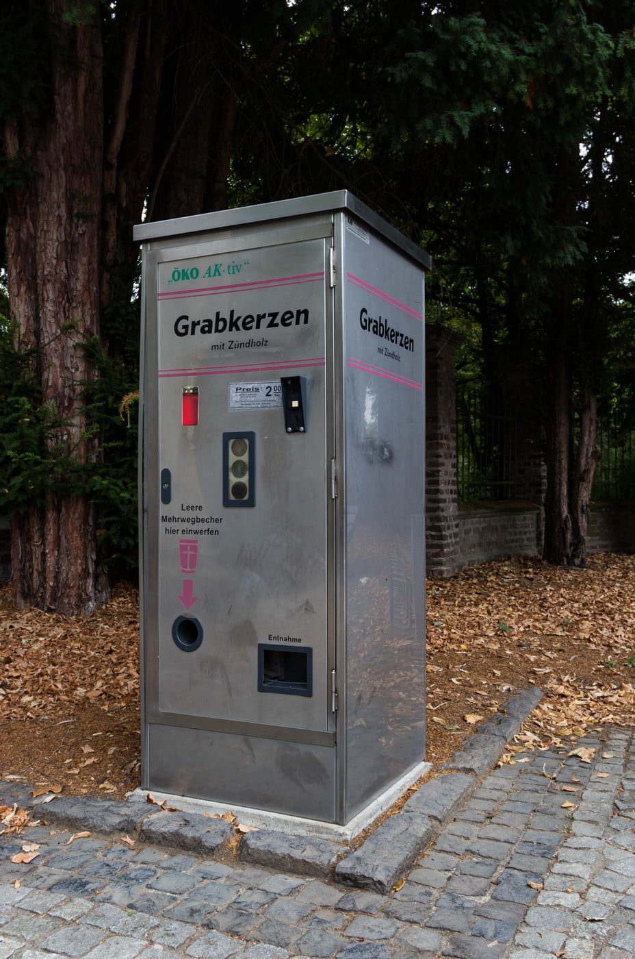 Grauer Verkaufsautomat für Grabkerzen auf dem Kopfsteinpflaster unter hohen Nadelbäumen auf Nordfriedhof Bonn
