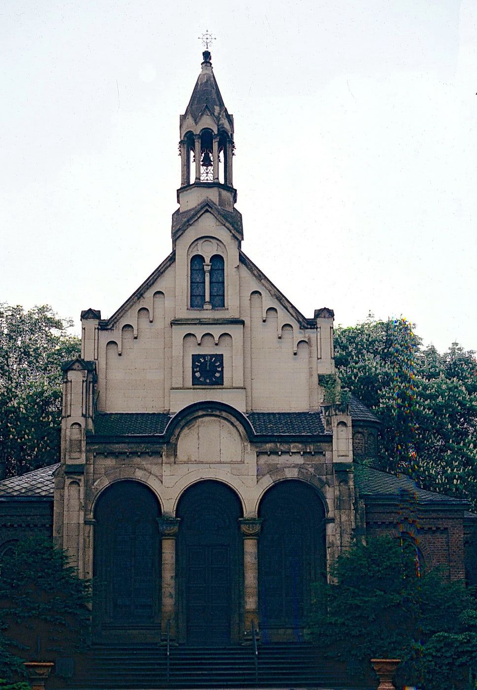Frontale historische Aufnahme der neuromanischen Kapelle mit dreibogigem Portal, Uhrgiebel und offenem Glockendachreiter. auf Nordfriedhof Bonn
